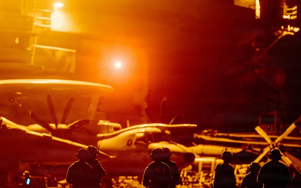 The image shows a naval aircraft carrier flight deck bathed in intense orange-amber light, likely from artificial deck lighting or a setting/rising sun. Multiple silhouetted personnel wearing helmets are visible on the deck alongside parked military aircraft. The dramatic lighting creates strong shadows and a high-contrast silhouette effect across the entire scene.