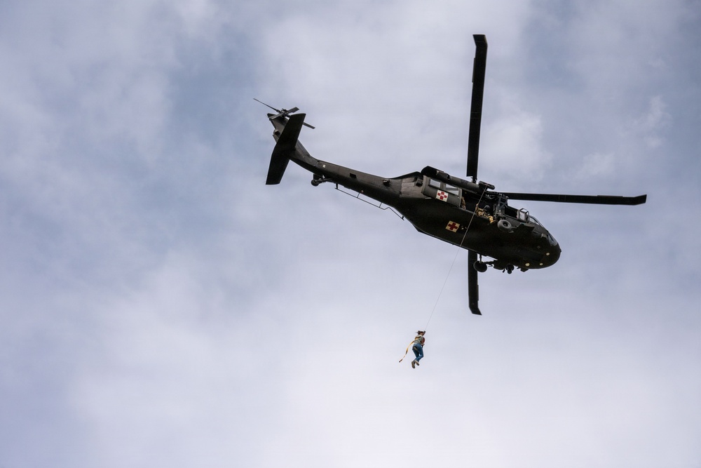 A dark olive/black military medical helicopter is captured in flight from a low-angle perspective against a partly cloudy sky. A person is suspended below the helicopter on a hoist or cable, indicating a medical evacuation or rescue operation. The helicopter displays red cross markings, identifying it as a medevac aircraft.