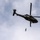 A dark olive/black military medical helicopter is captured in flight from a low-angle perspective against a partly cloudy sky. A person is suspended below the helicopter on a hoist or cable, indicating a medical evacuation or rescue operation. The helicopter displays red cross markings, identifying it as a medevac aircraft.