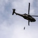 A dark olive/black military medical helicopter is captured in flight from a low-angle perspective against a partly cloudy sky. A person is suspended below the helicopter on a hoist or cable, indicating a medical evacuation or rescue operation. The helicopter displays red cross markings, identifying it as a medevac aircraft.