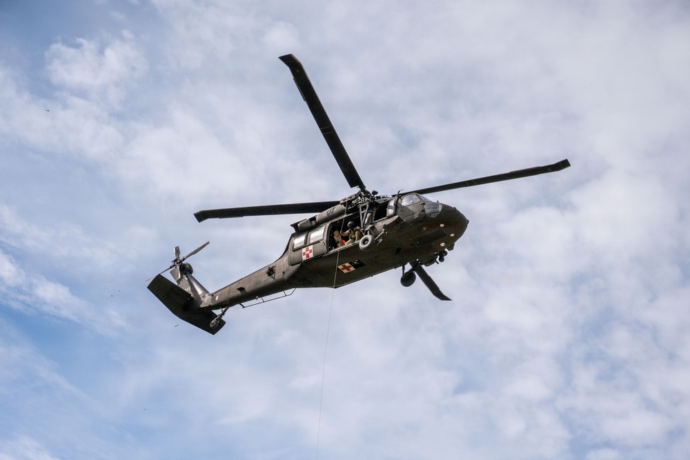 A military helicopter, identifiable as a UH-60 Black Hawk variant configured for medical evacuation (MEDEVAC), is photographed in flight from a low-angle perspective against a partly cloudy sky. The aircraft displays red cross markings indicating its medical role and has a hoist cable or rope visibly hanging from its underside. A crew member is visible in the open side door of the aircraft.