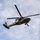 A military helicopter, identifiable as a UH-60 Black Hawk variant configured for medical evacuation (MEDEVAC), is photographed in flight from a low-angle perspective against a partly cloudy sky. The aircraft displays red cross markings indicating its medical role and has a hoist cable or rope visibly hanging from its underside. A crew member is visible in the open side door of the aircraft.