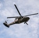 A military helicopter, identifiable as a UH-60 Black Hawk variant configured for medical evacuation (MEDEVAC), is photographed in flight from a low-angle perspective against a partly cloudy sky. The aircraft displays red cross markings indicating its medical role and has a hoist cable or rope visibly hanging from its underside. A crew member is visible in the open side door of the aircraft.