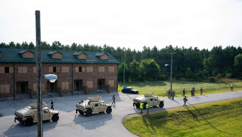 The image shows a military training exercise taking place at what appears to be a mock urban environment or training facility. Multiple HMMWV (Humvee) military vehicles are positioned along a road adjacent to a two-story brick building with boarded windows, suggesting a simulated urban operations scenario. Personnel in military uniforms and some in high-visibility vests are dispersed throughout the area.