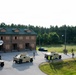 The image shows a military training exercise taking place at what appears to be a mock urban environment or training facility. Multiple HMMWV (Humvee) military vehicles are positioned along a road adjacent to a two-story brick building with boarded windows, suggesting a simulated urban operations scenario. Personnel in military uniforms and some in high-visibility vests are dispersed throughout the area.