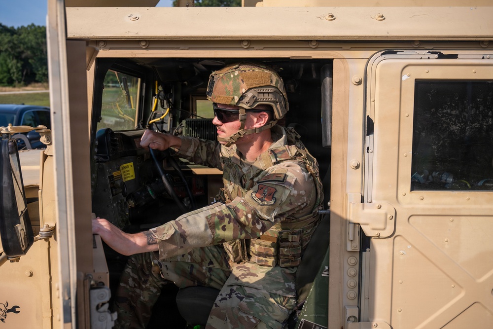 A military service member in full combat gear is seated in the driver's seat of a tan/desert-colored HMMWV (Humvee). The individual is wearing OCP (Operational Camouflage Pattern) uniform, a tactical helmet, ballistic sunglasses, and a plate carrier vest. The photo is taken from the open driver's side door, with a wooded outdoor environment visible in the background.