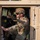 A military service member in full combat gear is seated in the driver's seat of a tan/desert-colored HMMWV (Humvee). The individual is wearing OCP (Operational Camouflage Pattern) uniform, a tactical helmet, ballistic sunglasses, and a plate carrier vest. The photo is taken from the open driver's side door, with a wooded outdoor environment visible in the background.