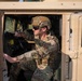 A military service member in full combat gear is seated in the driver's seat of a tan/desert-colored HMMWV (Humvee). The individual is wearing OCP (Operational Camouflage Pattern) uniform, a tactical helmet, ballistic sunglasses, and a plate carrier vest. The photo is taken from the open driver's side door, with a wooded outdoor environment visible in the background.