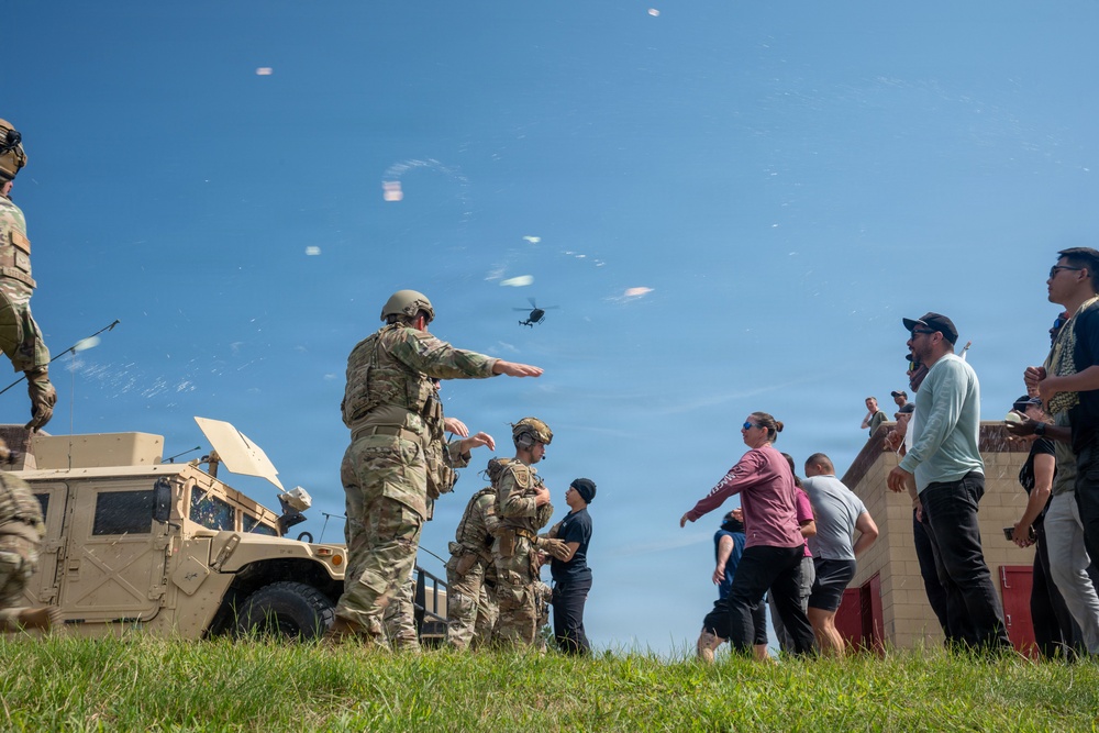 Military personnel in full combat gear interact with a group of civilians near a military HMMWV (Humvee) vehicle on a grassy field. A soldier appears to be directing or briefing the civilian group while a helicopter is visible in the background sky. The scene suggests a public demonstration, open house, or community engagement event at a military installation.