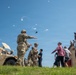 Military personnel in full combat gear interact with a group of civilians near a military HMMWV (Humvee) vehicle on a grassy field. A soldier appears to be directing or briefing the civilian group while a helicopter is visible in the background sky. The scene suggests a public demonstration, open house, or community engagement event at a military installation.