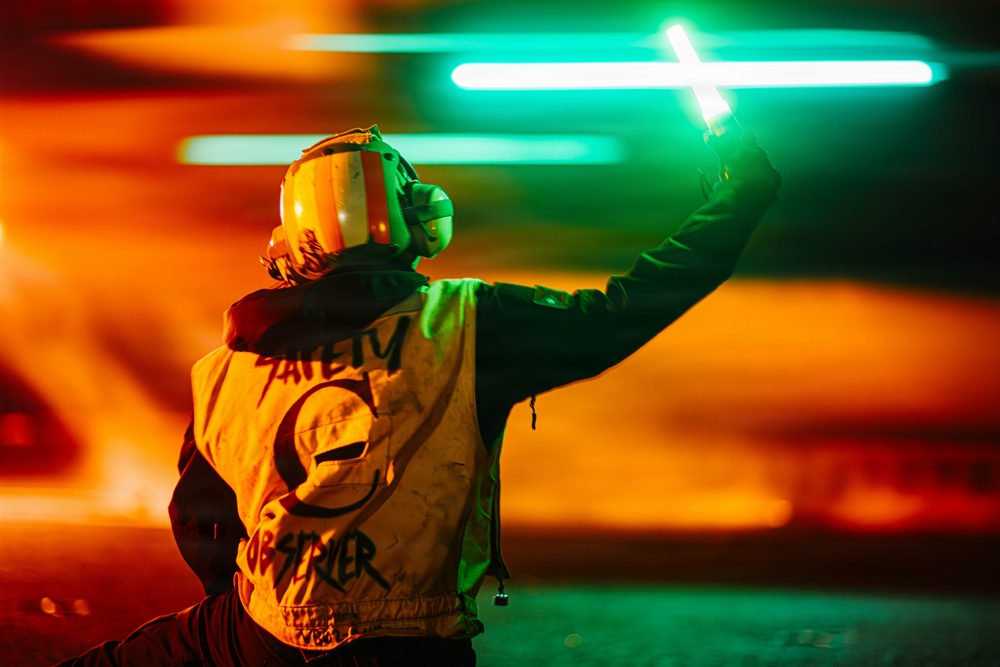 A person wearing a safety vest labeled 'Safety Observer' and a protective helmet with ear defenders is holding a glowing green light wand raised in the air. The image is taken at night with dramatic orange and teal/green lighting creating a vivid, high-contrast atmosphere. The scene appears to be on an airfield or similar operational environment where marshaling signals are used.