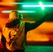 A person wearing a safety vest labeled 'Safety Observer' and a protective helmet with ear defenders is holding a glowing green light wand raised in the air. The image is taken at night with dramatic orange and teal/green lighting creating a vivid, high-contrast atmosphere. The scene appears to be on an airfield or similar operational environment where marshaling signals are used.