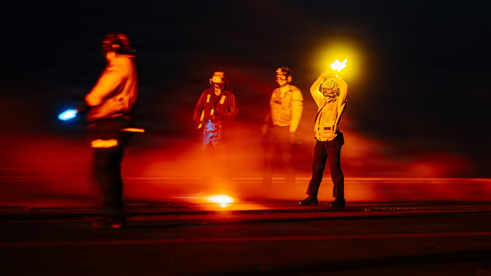 A nighttime scene on what appears to be an aircraft carrier flight deck, with several crew members visible under dramatic red and orange lighting. Personnel are wearing helmets, protective gear, and colored jerseys typical of carrier deck operations. One crew member is holding a raised illuminated wand/baton, consistent with aircraft handling or marshaling duties.