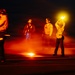 A nighttime scene on what appears to be an aircraft carrier flight deck, with several crew members visible under dramatic red and orange lighting. Personnel are wearing helmets, protective gear, and colored jerseys typical of carrier deck operations. One crew member is holding a raised illuminated wand/baton, consistent with aircraft handling or marshaling duties.