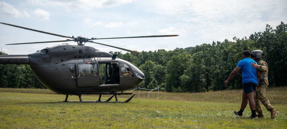 A military helicopter, appearing to be a UH-72 Lakota, is landed on a grassy field while a soldier in camouflage uniform and flight helmet escorts a civilian in a blue shirt toward the aircraft. The scene appears to take place during a training exercise, demonstration, or medical evacuation scenario in a rural, tree-lined area. The helicopter's door is open and a crew member is visible inside.