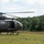 A military helicopter, appearing to be a UH-72 Lakota, is landed on a grassy field while a soldier in camouflage uniform and flight helmet escorts a civilian in a blue shirt toward the aircraft. The scene appears to take place during a training exercise, demonstration, or medical evacuation scenario in a rural, tree-lined area. The helicopter's door is open and a crew member is visible inside.