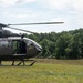 A military helicopter, appearing to be a UH-72 Lakota, is landed on a grassy field while a soldier in camouflage uniform and flight helmet escorts a civilian in a blue shirt toward the aircraft. The scene appears to take place during a training exercise, demonstration, or medical evacuation scenario in a rural, tree-lined area. The helicopter's door is open and a crew member is visible inside.