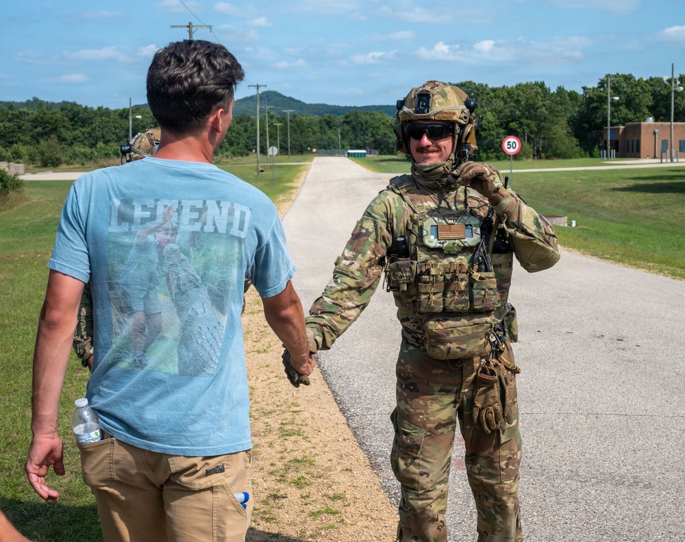 A fully equipped military operator in multicam camouflage gear, tactical vest, and helmet is shaking hands with a civilian man wearing a light blue 'LEGEND' t-shirt and khaki pants. The interaction appears friendly and takes place on a paved road at what looks like a military installation or training facility. The background shows a long straight road, green trees, a speed limit sign reading '50', and a brick building.
