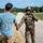 A fully equipped military operator in multicam camouflage gear, tactical vest, and helmet is shaking hands with a civilian man wearing a light blue 'LEGEND' t-shirt and khaki pants. The interaction appears friendly and takes place on a paved road at what looks like a military installation or training facility. The background shows a long straight road, green trees, a speed limit sign reading '50', and a brick building.