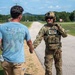 A fully equipped military operator in multicam camouflage gear, tactical vest, and helmet is shaking hands with a civilian man wearing a light blue 'LEGEND' t-shirt and khaki pants. The interaction appears friendly and takes place on a paved road at what looks like a military installation or training facility. The background shows a long straight road, green trees, a speed limit sign reading '50', and a brick building.
