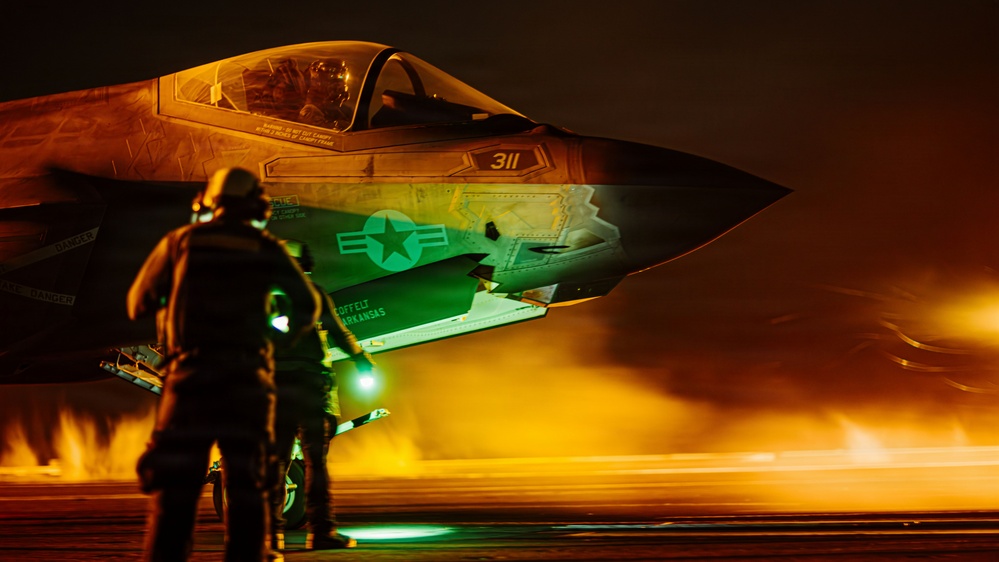 A night operations scene showing an F-35 stealth fighter jet (tail number 311) on what appears to be an aircraft carrier or airfield deck. Ground crew personnel are conducting pre-flight or post-flight checks under dramatic orange and green lighting. The aircraft displays U.S. military insignia and text referencing 'Arkansas' on its fuselage.