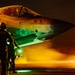 A night operations scene showing an F-35 stealth fighter jet (tail number 311) on what appears to be an aircraft carrier or airfield deck. Ground crew personnel are conducting pre-flight or post-flight checks under dramatic orange and green lighting. The aircraft displays U.S. military insignia and text referencing 'Arkansas' on its fuselage.