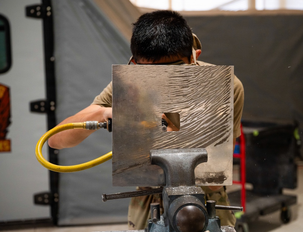 A person in military-style tan/olive clothing is working on a metal plate secured in a bench vise. They are using a pneumatic tool connected to a yellow air hose to work on a rectangular hole cut into the metal sheet. The metal plate shows distinctive grinding or sanding marks in a wave-like pattern across its surface.