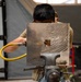 A person in military-style tan/olive clothing is working on a metal plate secured in a bench vise. They are using a pneumatic tool connected to a yellow air hose to work on a rectangular hole cut into the metal sheet. The metal plate shows distinctive grinding or sanding marks in a wave-like pattern across its surface.