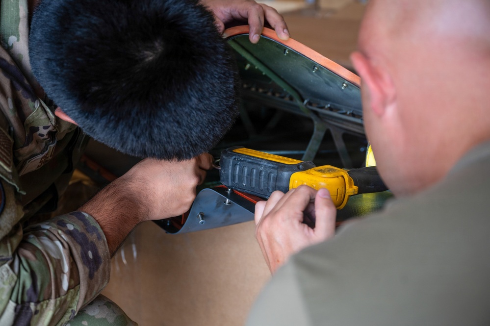 Two military personnel in uniform are working together on a mechanical or equipment assembly task. One individual wearing multicam/OCP camouflage uniform is using a yellow and black DeWalt power tool, while a second person in an olive/tan uniform assists. They appear to be focused on a component with orange and green elements, likely part of military equipment.
