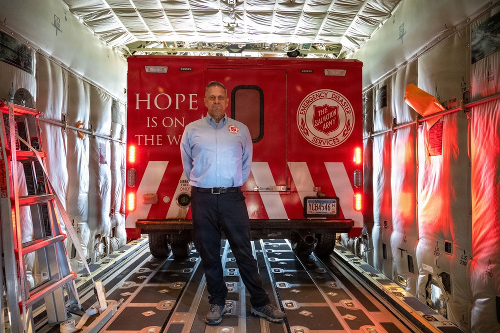 A man in a light blue shirt stands inside the cargo hold of a large aircraft in front of a red Salvation Army Emergency Disaster Services vehicle. The truck bears the text 'Hope Is On The Way' and the Salvation Army shield logo. The setting appears to be a military or cargo aircraft being used to transport disaster relief equipment.