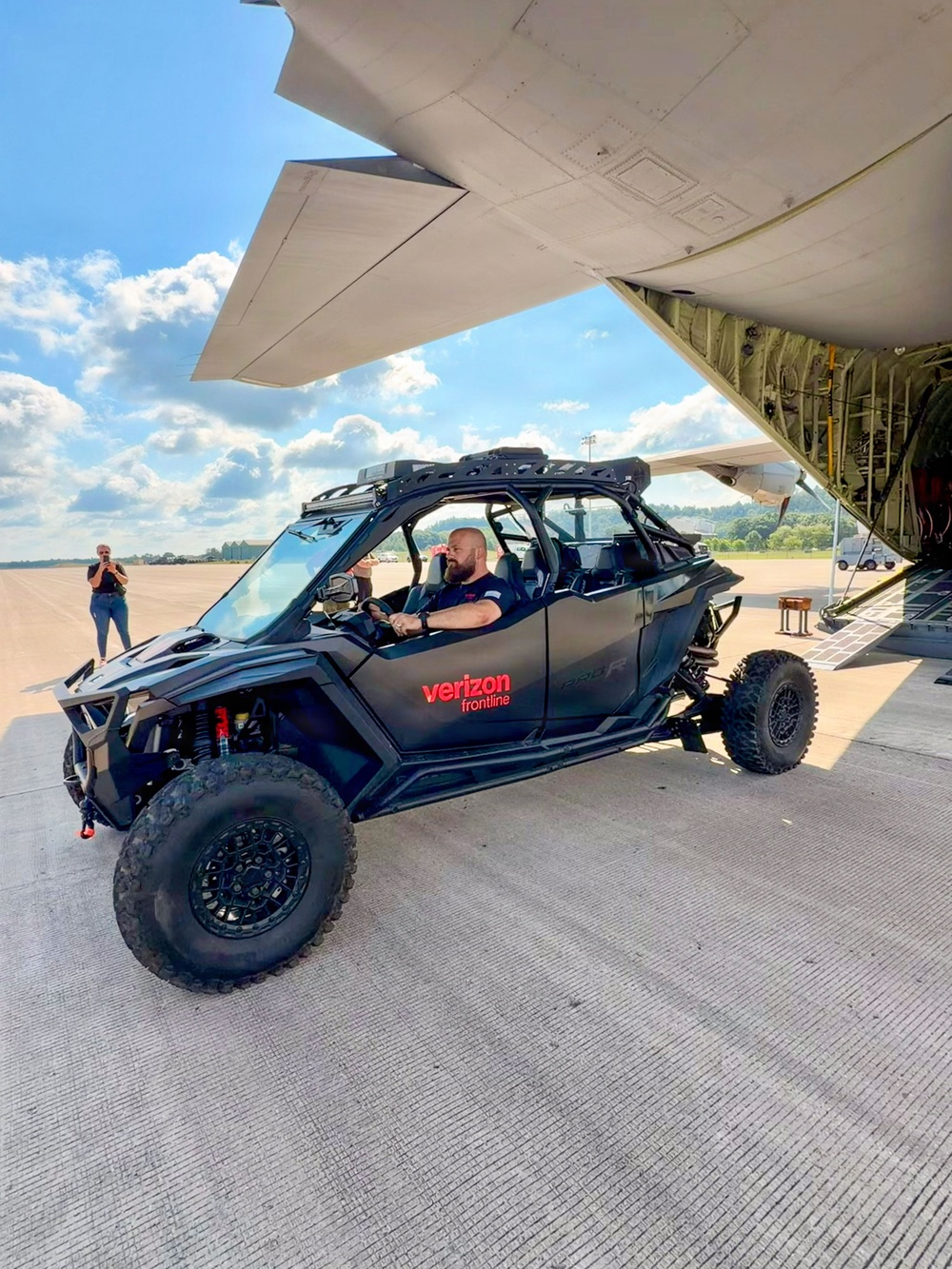 A black Verizon Frontline-branded off-road UTV (side-by-side vehicle) is being unloaded from a large military-style cargo aircraft on a tarmac. A bearded man is seated in the driver's seat, while another person stands in the background photographing the scene. The setting appears to be an airfield on a partly cloudy day, suggesting a disaster response or emergency deployment operation.
