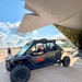 A black Verizon Frontline-branded off-road UTV (side-by-side vehicle) is being unloaded from a large military-style cargo aircraft on a tarmac. A bearded man is seated in the driver's seat, while another person stands in the background photographing the scene. The setting appears to be an airfield on a partly cloudy day, suggesting a disaster response or emergency deployment operation.