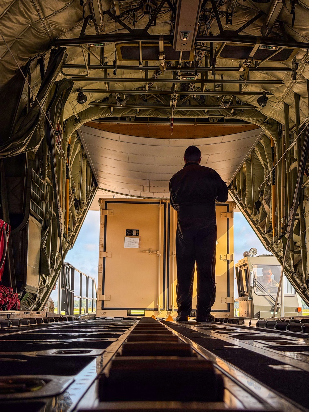 The image is taken from inside the cargo hold of a military transport aircraft, looking toward the open rear ramp. A person stands silhouetted against the bright exterior light, overseeing the loading of a large tan/beige military cargo container. A ground support vehicle with an operator is visible outside, positioned to load or unload the container.