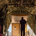 The image is taken from inside the cargo hold of a military transport aircraft, looking toward the open rear ramp. A person stands silhouetted against the bright exterior light, overseeing the loading of a large tan/beige military cargo container. A ground support vehicle with an operator is visible outside, positioned to load or unload the container.