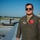 A male military aviator in an olive green flight suit stands on or near the wing of a military aircraft on a flight line. He is wearing aviator sunglasses and has multiple patches visible on his flight suit, including what appears to be a U.S. Air National Guard unit patch. In the background, at least one C-130 Hercules transport aircraft marked 'U.S. AIR FORCE' is visible on the tarmac.