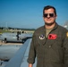 A male military aviator in an olive green flight suit stands on or near the wing of a military aircraft on a flight line. He is wearing aviator sunglasses and has multiple patches visible on his flight suit, including what appears to be a U.S. Air National Guard unit patch. In the background, at least one C-130 Hercules transport aircraft marked 'U.S. AIR FORCE' is visible on the tarmac.