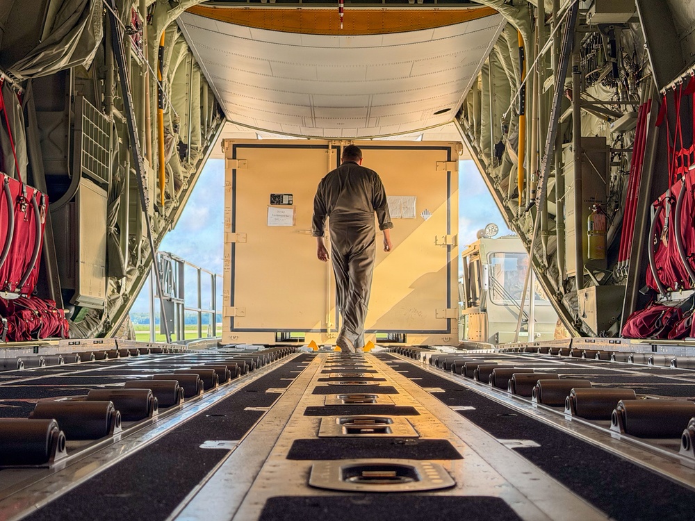 The image shows the interior of a military cargo aircraft, viewed from the rear looking toward the open aft ramp. A single person in gray flight suit or coveralls is walking toward a large pale-yellow cargo container positioned at the ramp opening. The scene appears to be during a loading or inspection operation on an airfield.
