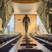 The image shows the interior of a military cargo aircraft, viewed from the rear looking toward the open aft ramp. A single person in gray flight suit or coveralls is walking toward a large pale-yellow cargo container positioned at the ramp opening. The scene appears to be during a loading or inspection operation on an airfield.