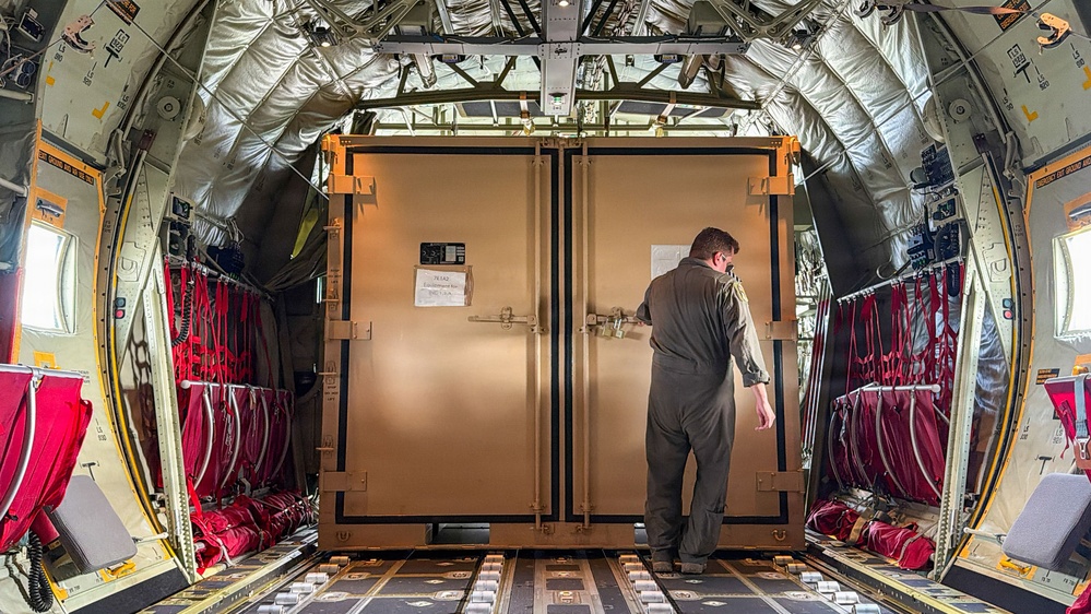 The interior of a military cargo aircraft shows a large tan/brown equipment container secured on the cargo floor. A person wearing a military flight suit is standing at the container's double doors, appearing to inspect or access it. The aircraft interior features characteristic insulated walls, red cargo straps/nets, and a roller-equipped cargo floor with tie-down tracks.