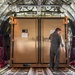 The interior of a military cargo aircraft shows a large tan/brown equipment container secured on the cargo floor. A person wearing a military flight suit is standing at the container's double doors, appearing to inspect or access it. The aircraft interior features characteristic insulated walls, red cargo straps/nets, and a roller-equipped cargo floor with tie-down tracks.