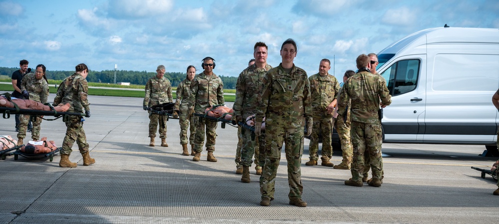A group of military personnel in camouflage uniforms are conducting what appears to be a medical evacuation or casualty handling exercise on an airfield tarmac. Several soldiers are carrying stretchers with training mannequins/simulated casualties, while others observe or participate in the drill. A white cargo van is parked nearby, likely serving as a support vehicle for the exercise.