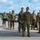A group of military personnel in camouflage uniforms are conducting what appears to be a medical evacuation or casualty handling exercise on an airfield tarmac. Several soldiers are carrying stretchers with training mannequins/simulated casualties, while others observe or participate in the drill. A white cargo van is parked nearby, likely serving as a support vehicle for the exercise.
