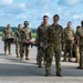 A group of military personnel in camouflage uniforms are conducting what appears to be a medical evacuation or casualty handling exercise on an airfield tarmac. Several soldiers are carrying stretchers with training mannequins/simulated casualties, while others observe or participate in the drill. A white cargo van is parked nearby, likely serving as a support vehicle for the exercise.