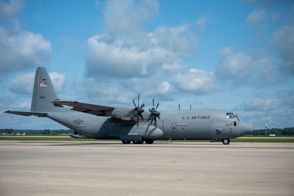 A U.S. Air Force C-130J Super Hercules military transport aircraft is parked on a tarmac under a partly cloudy blue sky. The aircraft is painted in standard gray livery and displays visible markings including 'U.S. AIR FORCE' on the fuselage, an American flag on the tail, and the serial number 16003. The setting appears to be a military airfield with a tree line visible in the background.