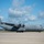 A U.S. Air Force C-130J Super Hercules military transport aircraft is parked on a tarmac under a partly cloudy blue sky. The aircraft is painted in standard gray livery and displays visible markings including 'U.S. AIR FORCE' on the fuselage, an American flag on the tail, and the serial number 16003. The setting appears to be a military airfield with a tree line visible in the background.