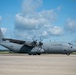 A U.S. Air Force C-130J Super Hercules military transport aircraft is parked on a tarmac under a partly cloudy blue sky. The aircraft is painted in standard gray livery and displays visible markings including 'U.S. AIR FORCE' on the fuselage, an American flag on the tail, and the serial number 16003. The setting appears to be a military airfield with a tree line visible in the background.