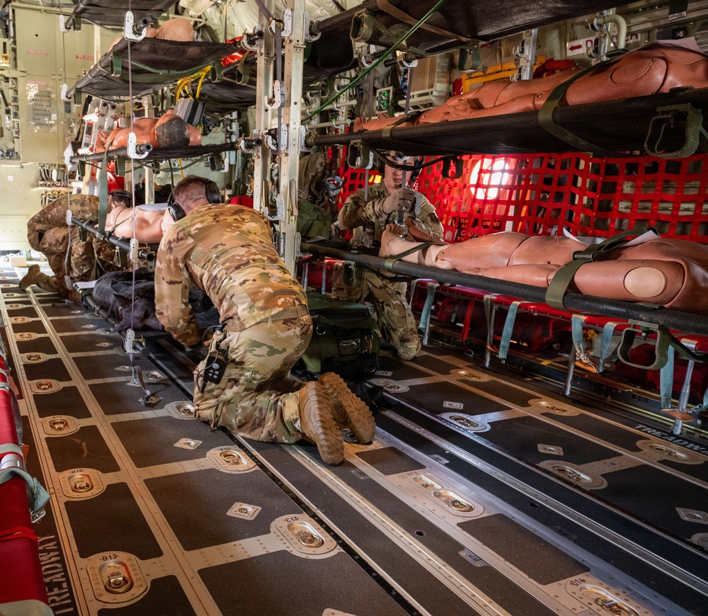 The image shows the interior of a military transport aircraft configured for aeromedical evacuation, with multiple litter (stretcher) stanchion systems loaded with medical training mannequins. At least two military personnel in OCP (Operational Camouflage Pattern) uniforms are working on the floor and near the litter systems, appearing to conduct a training exercise. The aircraft interior features a red cargo net partition, treadway floor panels with tie-down rings, and medical equipment mounted along the fuselage walls.