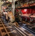 The image shows the interior of a military transport aircraft configured for aeromedical evacuation, with multiple litter (stretcher) stanchion systems loaded with medical training mannequins. At least two military personnel in OCP (Operational Camouflage Pattern) uniforms are working on the floor and near the litter systems, appearing to conduct a training exercise. The aircraft interior features a red cargo net partition, treadway floor panels with tie-down rings, and medical equipment mounted along the fuselage walls.
