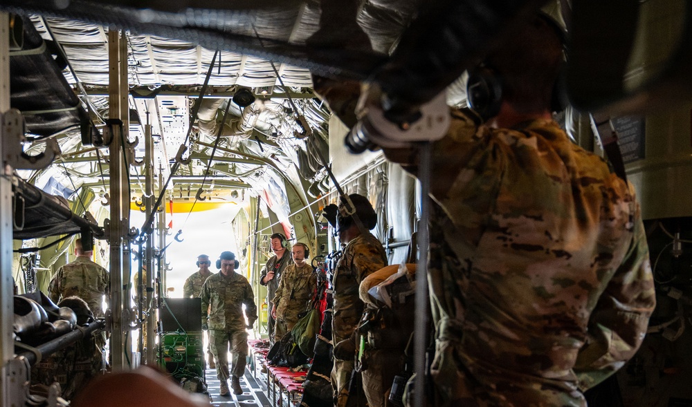 Military personnel in MultiCam/OCP camouflage uniforms are gathered inside the cargo hold of a large military transport aircraft, likely a C-130 Hercules. Several soldiers wearing communications headsets are visible, with the rear cargo ramp open, allowing natural light to illuminate the interior. The scene appears to depict a pre-mission briefing, loading, or aeromedical evacuation preparation.