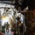 Military personnel in MultiCam/OCP camouflage uniforms are gathered inside the cargo hold of a large military transport aircraft, likely a C-130 Hercules. Several soldiers wearing communications headsets are visible, with the rear cargo ramp open, allowing natural light to illuminate the interior. The scene appears to depict a pre-mission briefing, loading, or aeromedical evacuation preparation.